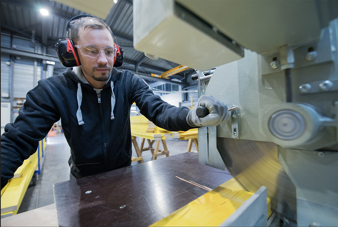 un compagnon qui coupe à l'aire d'une machine à bois. Il porte des protections auditives.